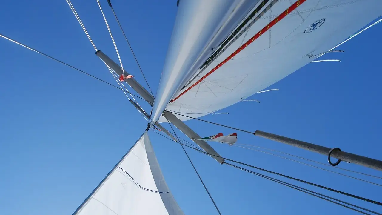 Vue plongeante sur le mat de bateau, voiles blanches déployées sous un ciel bleu éclatant, cordages tendus.