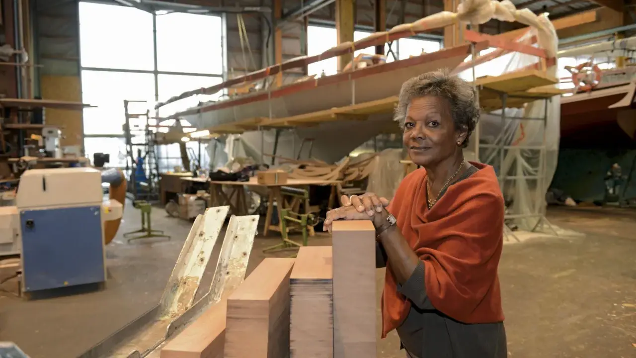 Jacqueline Tabarly, souriante, dans un atelier de construction navale, devant des blocs de bois et un voilier en cours de fabrication.