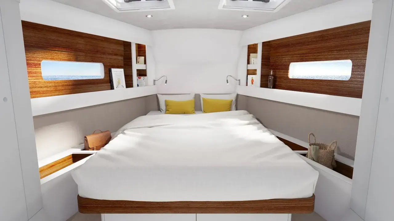 L'intérieur d'un voilier 8m, avec un grand lit blanc, des coussins jaunes, des hublots donnant sur la mer et des étagères en bois.