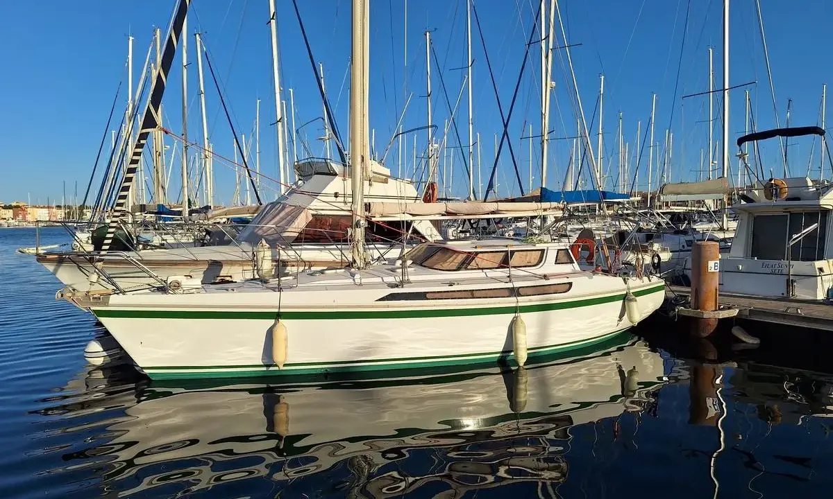 Un voilier blanc et vert, un Jeanneau Espace 1000, reflète sur l'eau calme d'un port.