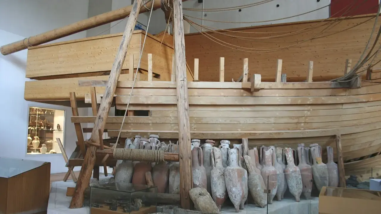 Amphores antiques alignées devant une reconstitution de navire en bois, rappelant le transport de vin par un ancien pinardier.