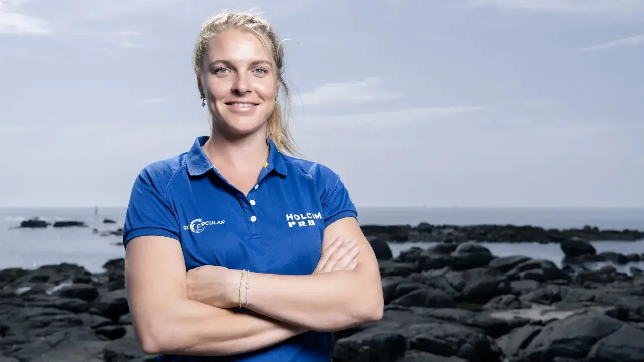 Rosalin Kuiper, souriante, pose les bras croisés devant un paysage côtier rocheux.