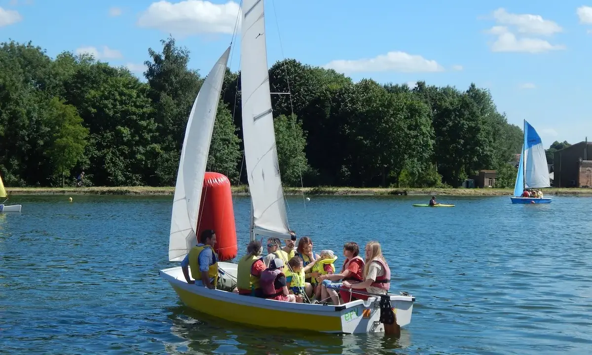Un groupe de personnes, dont des enfants, profite d'une journée ensoleillée sur un bouchain bateau jaune. Un autre bateau à voile bleu navigue au loin.