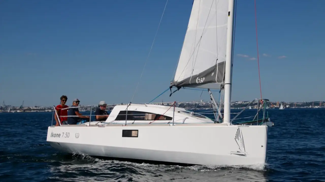 Un voilier Ikone 7.50 navigue par beau temps, avec trois personnes à bord. La voile est hissée, le ciel est bleu.