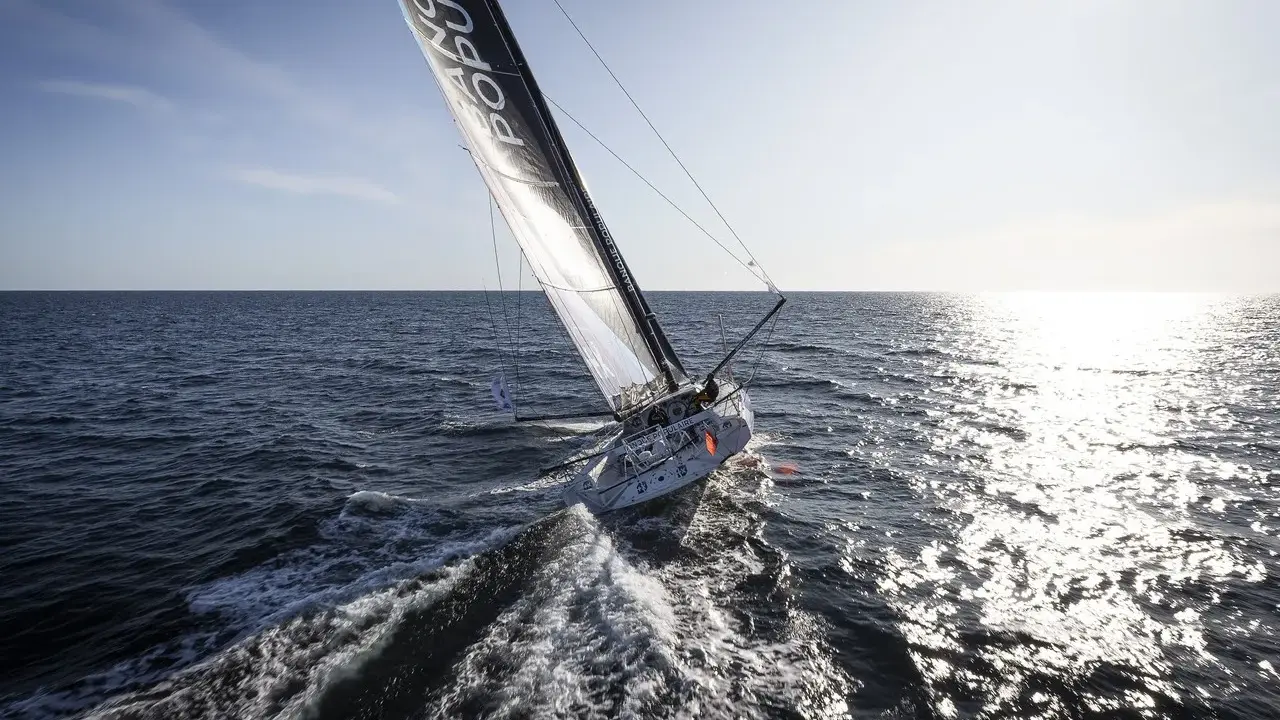 Voilier Banque Populaire naviguant au soleil, l'eau éclabousse le long du babord tribord.