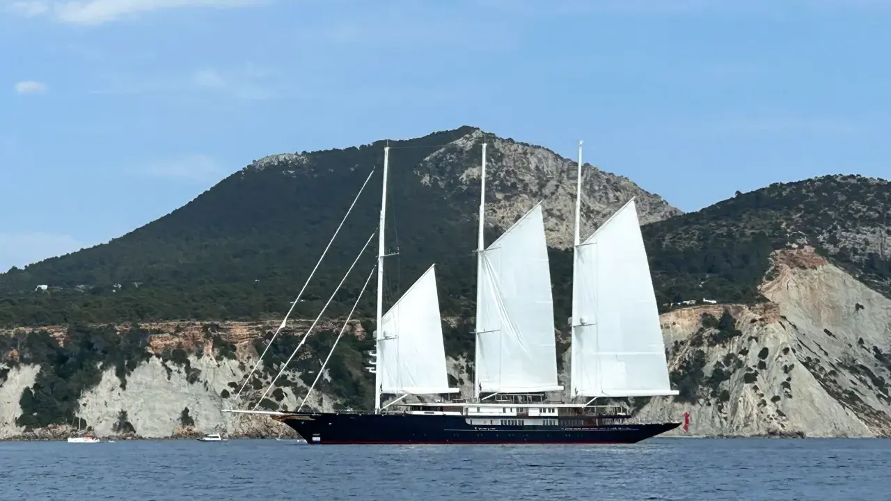 Le yacht de Bezos, un voilier majestueux, navigue en Méditerranée, ses voiles blanches déployées devant des falaises escarpées.