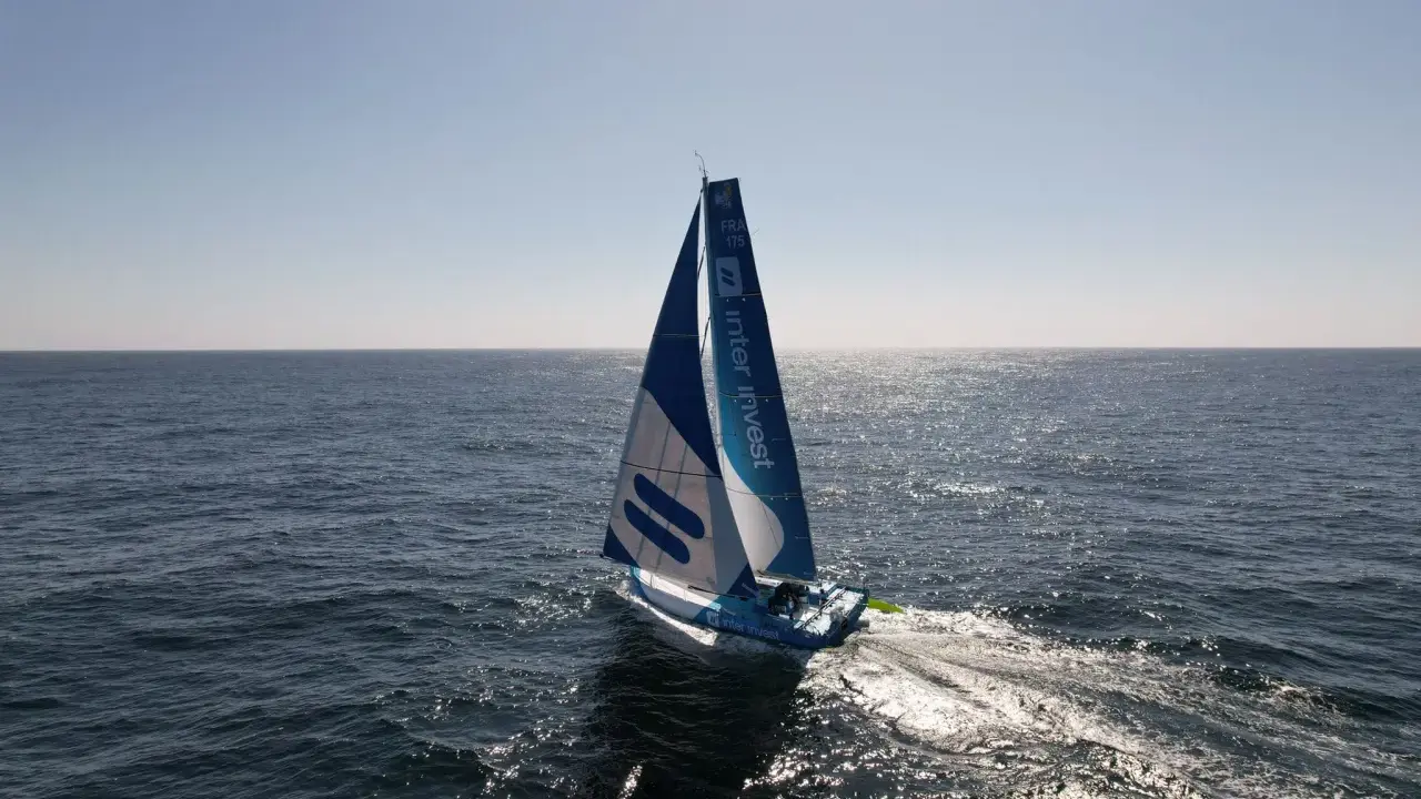 Voilier bleu et blanc "Inter Invest" naviguant sur une mer calme. Matthieu Perraut, skipper, est à la barre.
