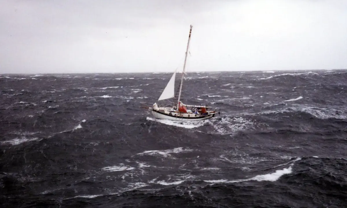Un voilier blanc lutte contre les vagues déchaînées dans une mer agitée. Le ciel est gris et menaçant, annonçant une tempête.