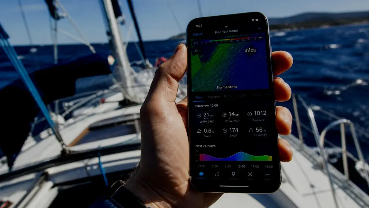 Sur un voilier, une main tient un smartphone affichant une application météo marine gratuite avec les prévisions pour Ibiza.