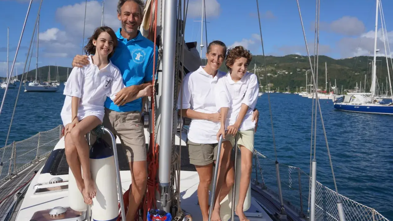Famille souriante sur un voilier, un moment clé de leur récit de voyage en voilier. La mer bleue et les voiliers en arrière-plan complètent ce tableau idyllique.