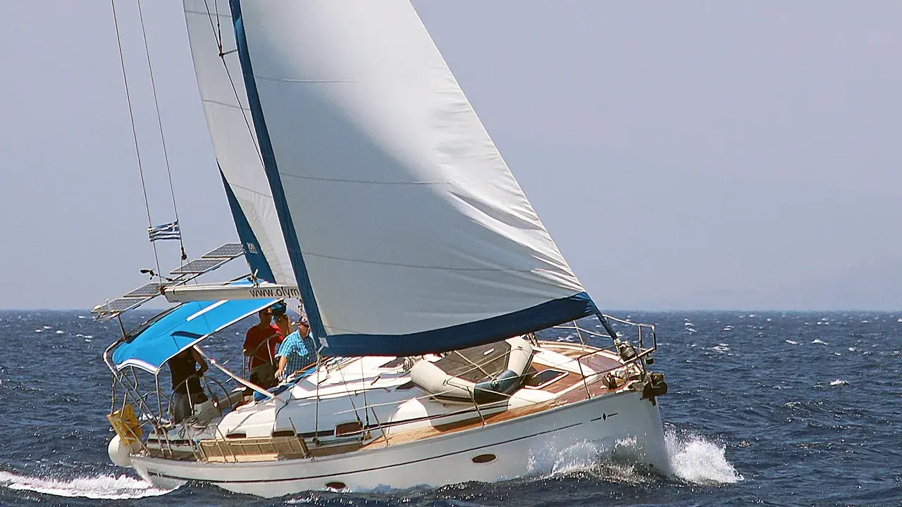 Un voilier blanc navigue sur une mer bleue agitée. L'équipage semble prendre un ris, ajustant les voiles pour mieux affronter le vent.