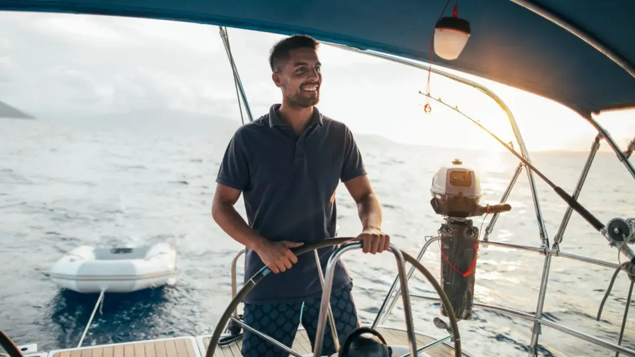 Un homme souriant barre un bateau, profitant du soleil couchant. Un petit bateau gonflable est attaché à l'arrière.