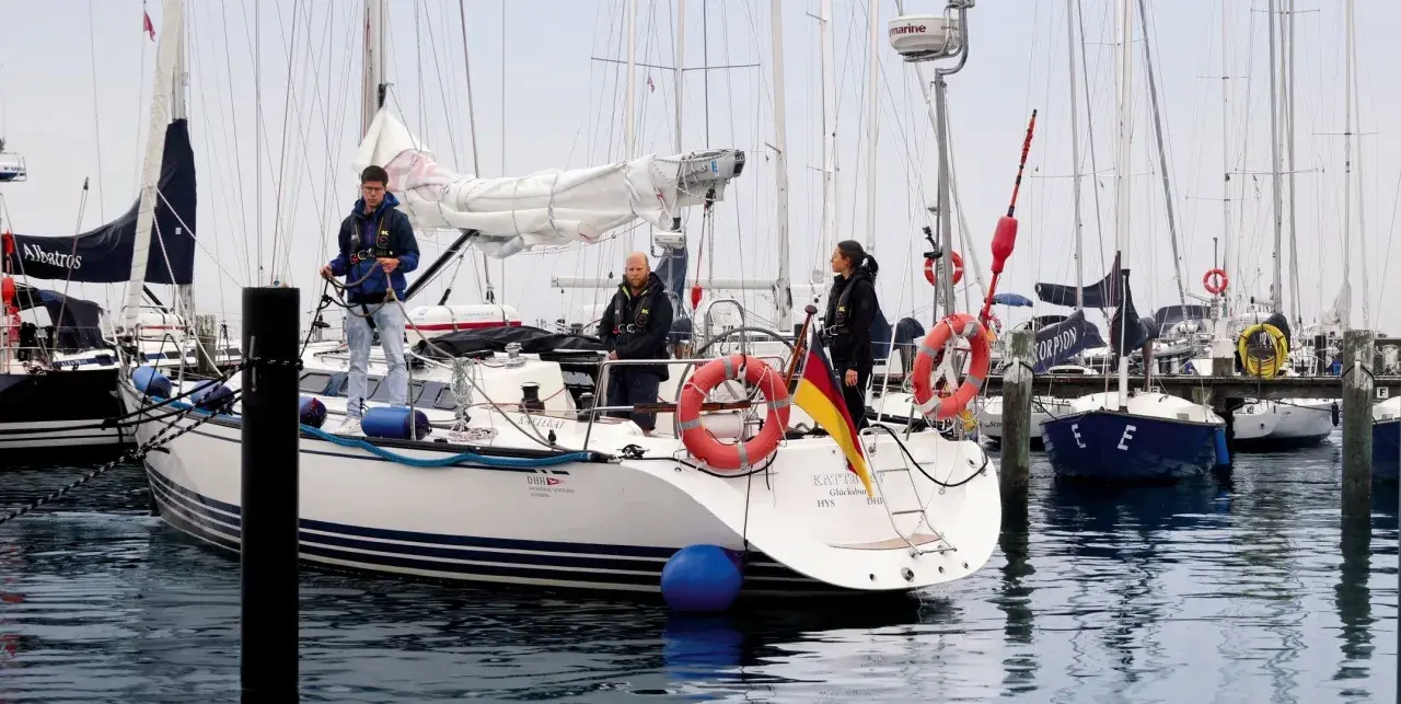 Trois personnes sur un bateau, prêtes à appareiller. Le drapeau allemand flotte fièrement. L'erre bateau est calme.