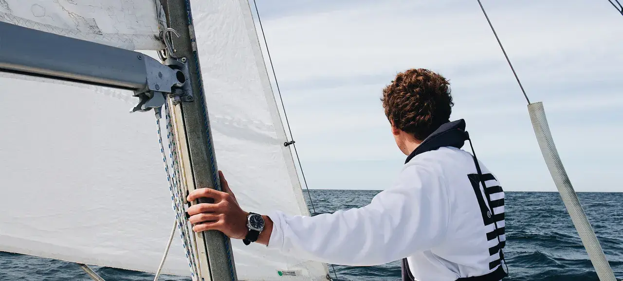 Un jeune homme regarde l'horizon depuis un voilier autonome. La voile blanche est gonflée par le vent.