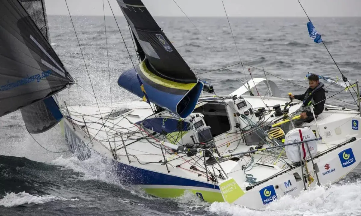 Un skipper navigue sur un voilier blanc et bleu, affrontant les vagues. Le nom "Pierre Quiroga" est visible sur la coque.