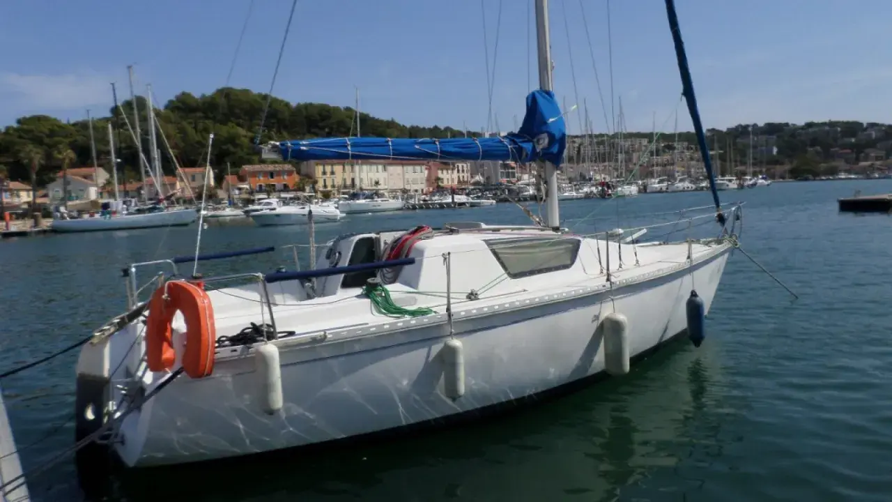 Bateau blanc à quai, voiles bleues enroulées. Au loin, un port animé avec des maisons colorées et d'autres voiliers. Un endroit idéal pour un séjour, comme le suggèrent les gib sea 76 avis.