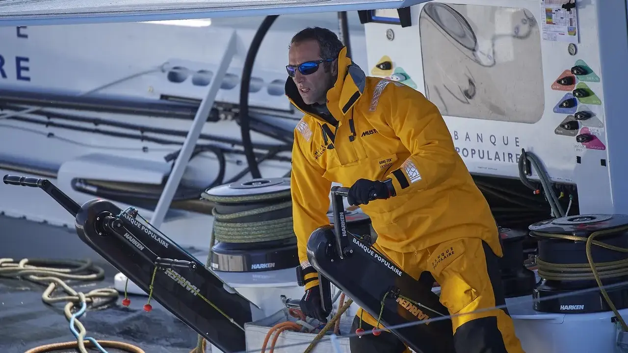 Skipper en tenue jaune, concentré, manœuvre un winch sur un bateau Banque Populaire.