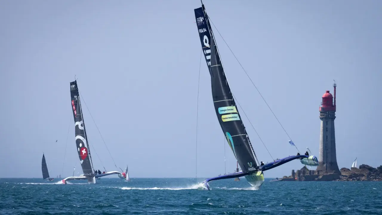 Course d'Ocean Fifty près d'un phare. Les trimarans filent sur l'eau, leurs voiles noires portant des logos.