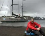 Mike Horn, l'explorateur, pose près de son bateau "Mercedes-Benz" sur une plage rocheuse, avec des montagnes enneigées en arrière-plan.