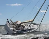 Un voilier échoué, incliné sur le côté, témoigne d'une possible collision bateau. Les vagues s'écrasent sur sa coque blanche sous un ciel bleu.