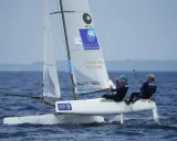 Deux marins, dont Billy Besson, naviguent sur un catamaran blanc par temps calme.