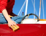 Nettoyage coque bateau vinaigre blanc : une main frotte la coque rouge avec une éponge savonneuse, sous un ciel bleu éclatant.