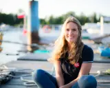 Irina Gracheva, souriante, assise sur un ponton en bois, avec des bateaux et des bouées en arrière-plan.