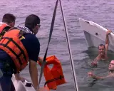 Sauvetage après un naufrage de bateau. Des secouristes en gilets orange aident des hommes dans l'eau, près d'une embarcation renversée.