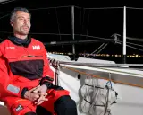 Giancarlo Pedote, skipper en tenue de voile rouge, sur un bateau la nuit, avec des lumières de ville en arrière-plan.
