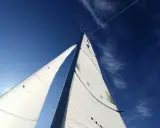 Voiles blanches gonflées par le vent, le bateau semble s'affaler sur l'eau bleue sous un ciel clair.