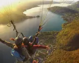 Vol en parapente biplace au-dessus d'un lac, avec un paysage de montagne et de village. Philippe Jeantot, malgré un accident passé, continue de voler.