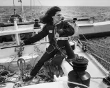 Une jeune femme aux cheveux longs, vêtue d'une combinaison de marin, manœuvre un winch sur un voilier. L'image évoque l'esprit d'aventure de Jacques Arthaud.