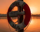 Bouée de sauvetage rouge et blanche sur un poteau, au bord d'une voie d'eau calme, avec un bateau à l'horizon au coucher du soleil.