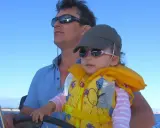 Un homme, le chef de bord bateau, tient son enfant sur ses genoux, naviguant sous un ciel bleu.