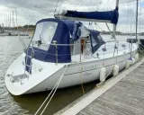 Un voilier blanc, un Océanis 311 Clipper, est amarré à un quai en bois. Des pare-battages blancs protègent sa coque.
