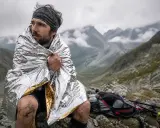 Un coureur transi, enveloppé dans une couverture de survie, contemple le paysage montagneux. Son sac à dos et ses bâtons de randonnée sont posés à côté.