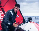 Gaston Morvan, marin concentré, ajuste une corde sur un voilier par temps couvert. La voile rouge porte des inscriptions blanches.