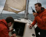 Deux hommes tentent de réparer un voilier en pleine mer. L'un utilise une lampe torche pour inspecter le moteur, tandis que l'autre communique par radio. Un **accident voilier** semble avoir eu lieu.