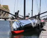 L'IMOCA Charal, un voilier de course futuriste, est amarré à quai. Des marins s'affairent sur le pont, préparant le navire pour une nouvelle aventure en mer.