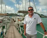 Loïc Bonnet, un homme souriant, pose sur un ponton bordé de voiliers. Il porte un polo Dream Yacht Charter, prêt pour une aventure en mer.