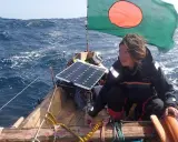 Capucine Trochet navigue sur une mer agitée, le drapeau du Bangladesh flottant au vent. Un panneau solaire est visible sur le bateau.