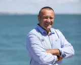 Lalou Roucayrol, souriant, bras croisés, devant la mer. Il porte une chemise bleue rayée avec un logo sur la poitrine.