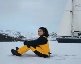 Tamara Klink, en tenue de ski jaune et noire, assise sur la neige, avec un voilier et des montagnes enneigées en arrière-plan.