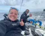 Hugo Dhallenne et sa coéquipière souriants sur un voilier, pouces levés, en pleine mer sous un ciel couvert.