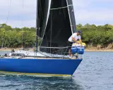 Un homme prépare le bateau, une voile noire déployée, sur fond de côte boisée. Il semble être le **half toner** de l'équipage.