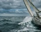 Un voilier fend les vagues sous un ciel menaçant. La proue découpe l'écume, le vent gonfle les voiles.