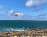 Un hélicoptère jaune et rouge survole une mer agitée, cherchant une personne disparue en mer. Un petit bateau rouge est visible au loin.