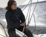 Marie Tabarly, souriante, tient la barre d'un voilier, le vent dans ses cheveux, naviguant sur une mer agitée.