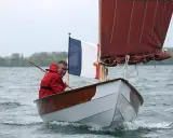 Un homme en ciré rouge navigue sur son bateau, fier du résultat de son travail pour construire son bateau. Le drapeau français flotte fièrement.