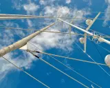 Vue plongeante sur le gréement d'un bateau, mâts blancs et cordages fins se découpant sur un ciel bleu et nuageux.